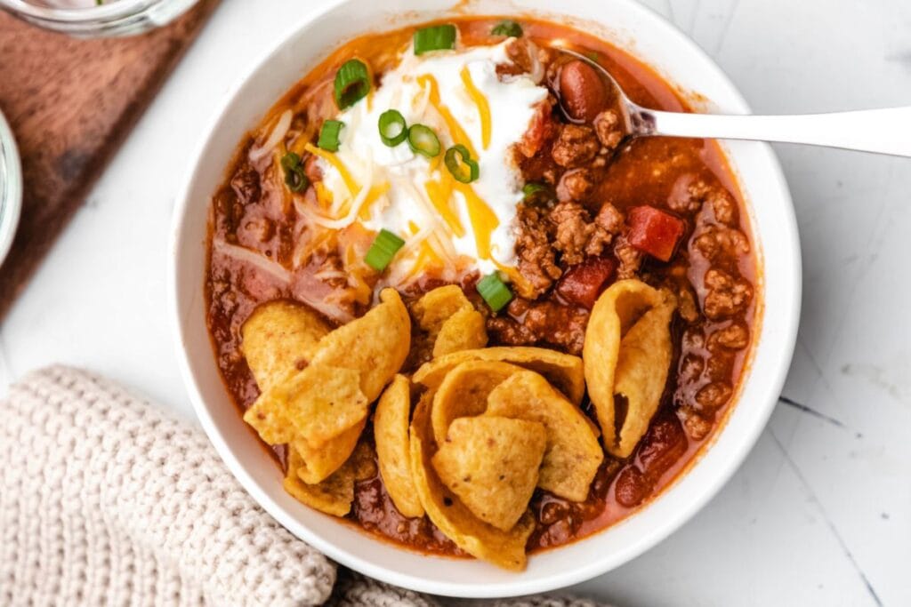 A bowl of chili topped with shredded cheese, sour cream, and chopped green onions. Several Fritos corn chips are also in the bowl.