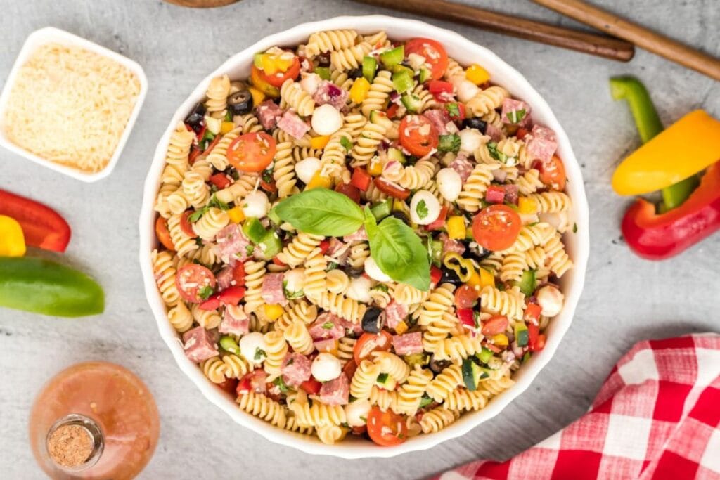 A white bowl of pasta salad with rotini pasta, salami, mozzarella balls, cherry tomatoes, bell peppers, and olives. A small bowl of grated cheese and sliced bell peppers are in the background.
