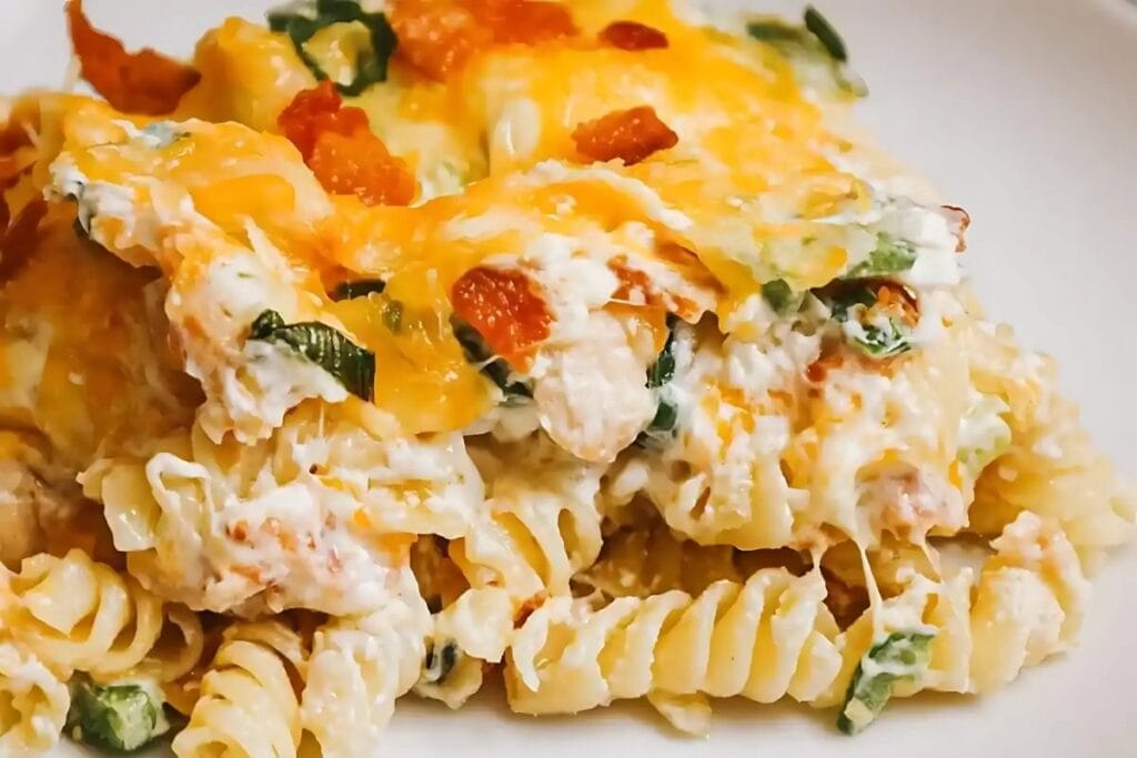 Close-up of a cheesy pasta dish. The pasta appears to be rotini, and the sauce is creamy with bits of green and red. The top is covered with melted cheese.