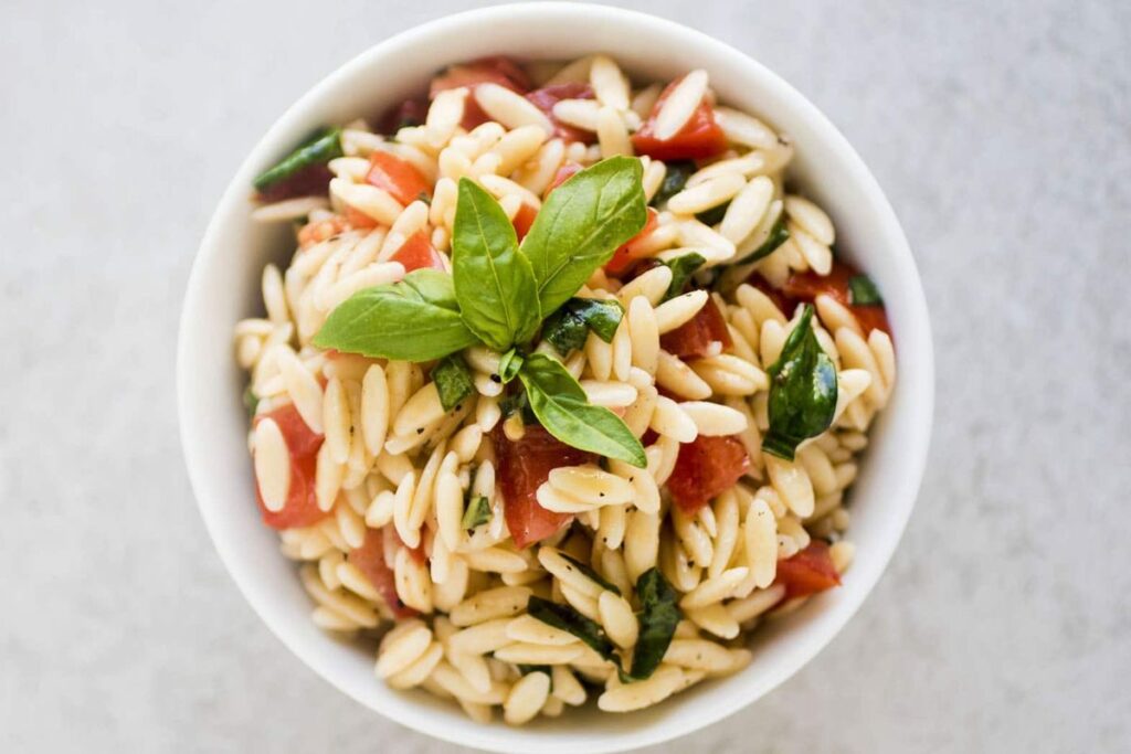 A white bowl of orzo pasta salad with diced tomatoes and fresh basil.