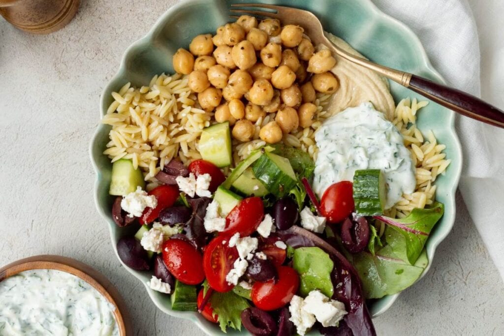 A bowl of Mediterranean-style food is shown from above. The bowl contains orzo pasta, chickpeas, a Greek salad with tomatoes, cucumbers, olives, and feta cheese, hummus, and tzatziki sauce. A small bowl of tzatziki sauce is visible in the lower left corner.