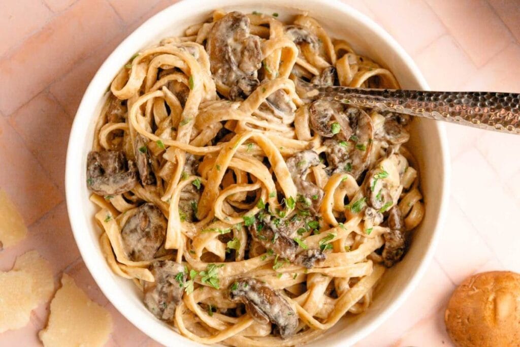 A bowl of creamy mushroom pasta is shown from above. The pasta is fettuccine, and the sauce is a creamy white sauce. Fresh parsley is sprinkled on top.