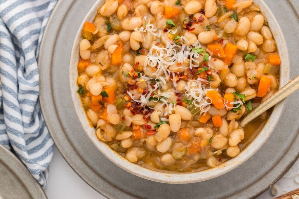 A bowl of creamy white bean soup is garnished with grated cheese, chopped parsley, and red pepper flakes. The bowl sits on a larger gray plate, and a blue and white striped napkin is partially visible in the background.