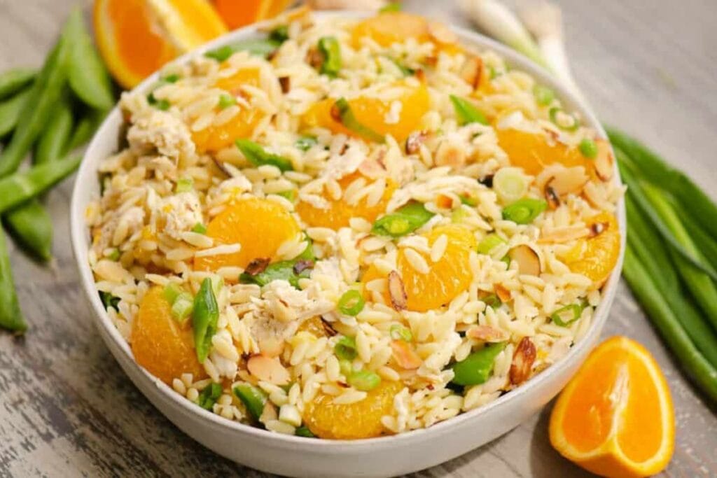 A bowl of orzo pasta salad with chicken, mandarin oranges, green peas, sliced almonds, and green onions.
