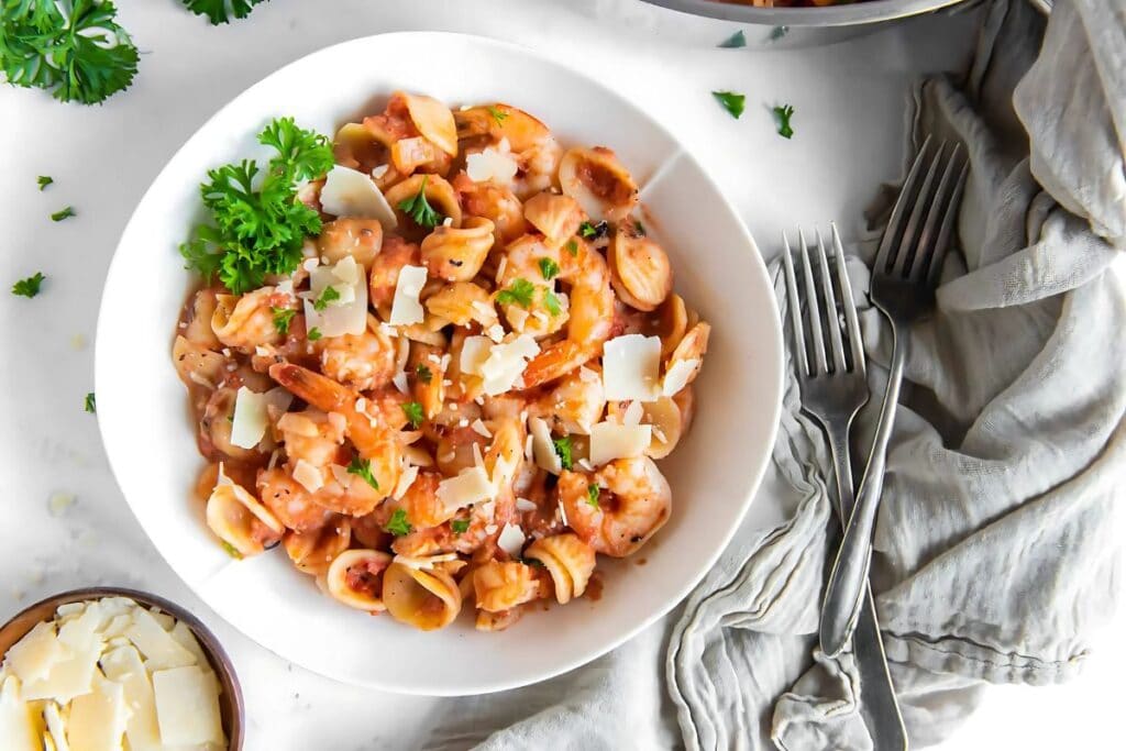 A white bowl of orecchiette pasta with shrimp in a vodka sauce is shown from above. The pasta is topped with grated Parmesan cheese and fresh parsley. Two forks and a small bowl of grated Parmesan cheese are visible in the background.