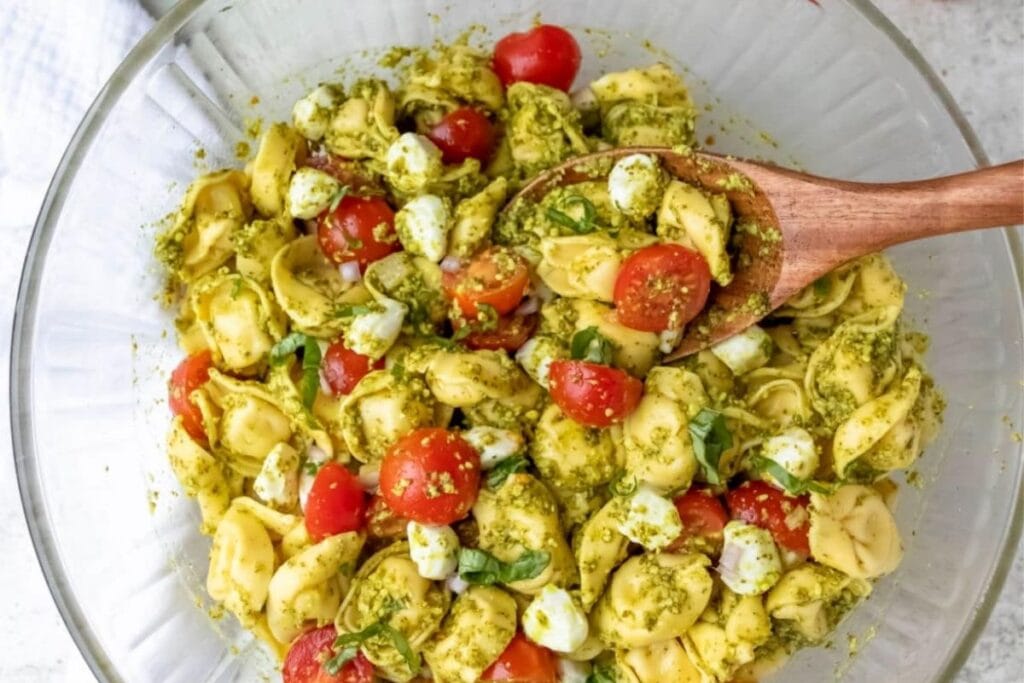 A glass bowl of tortellini pasta salad with pesto, cherry tomatoes, and mozzarella balls. A wooden spoon is in the bowl.