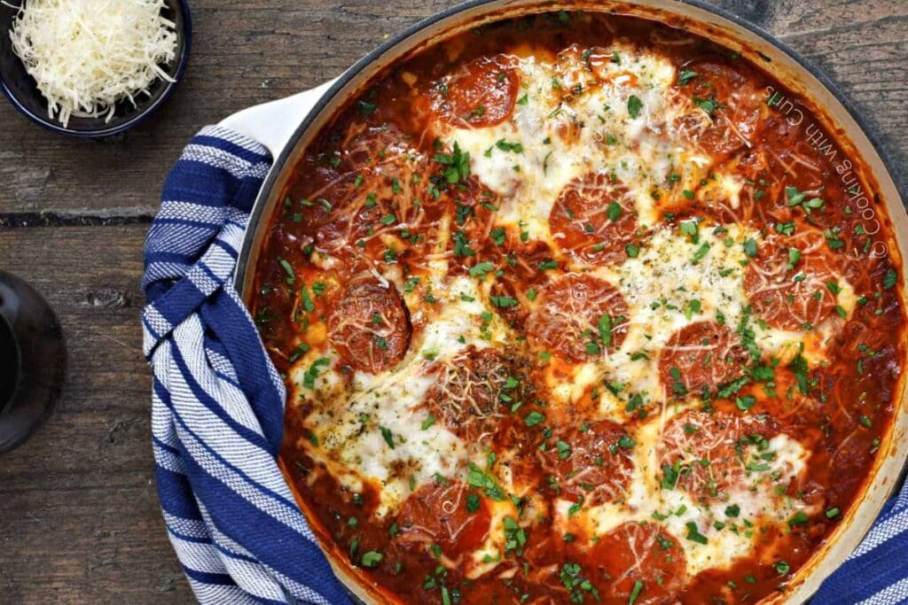 A large skillet of pizza chicken. The chicken is covered in tomato sauce, pepperoni, and melted cheese. Fresh parsley is sprinkled on top. A small bowl of shredded cheese is visible in the upper left corner.