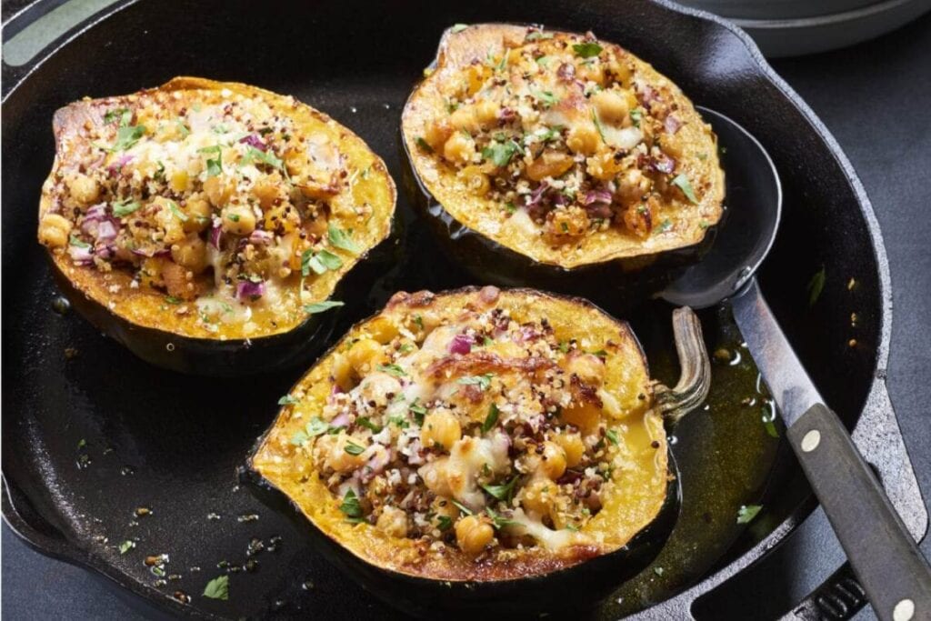 Three acorn squash halves are filled with a quinoa and chickpea mixture, topped with cheese, and baked in a cast iron skillet. A serving spoon is in the skillet.