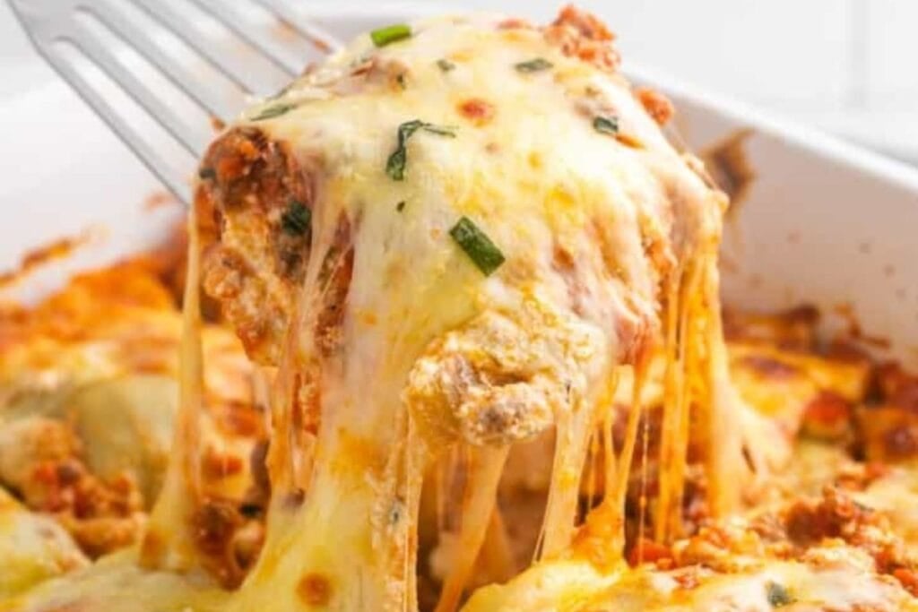 A serving utensil lifts a portion of baked ravioli lasagna. The lasagna is covered in melted cheese and is slightly browned on top. A bit of green garnish is visible.