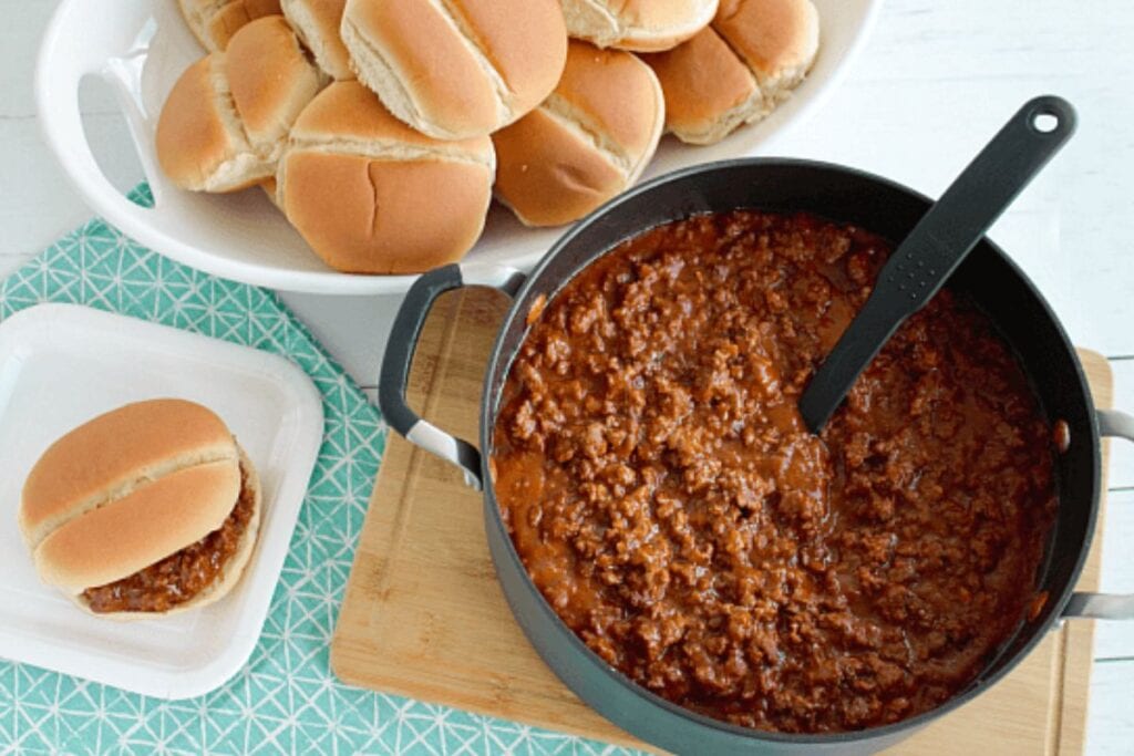 A large skillet of sloppy joe meat is shown with a serving spoon. A bowl of slider buns and a small plate with a sloppy joe slider are also visible.