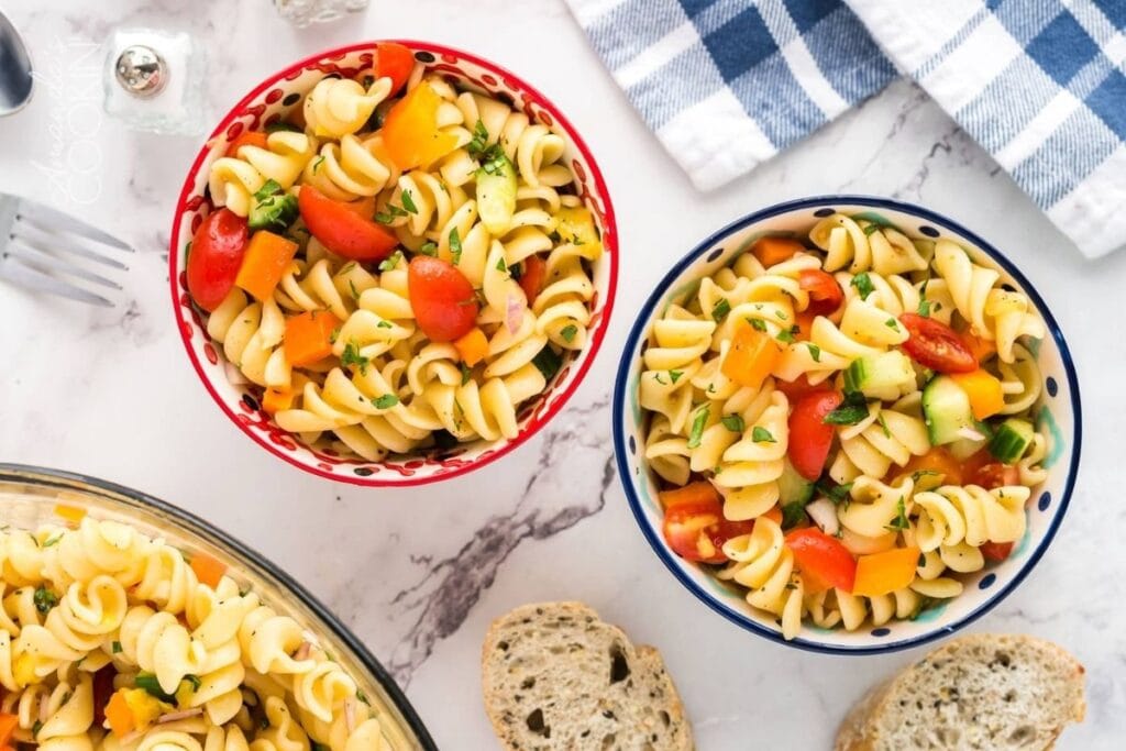 Two small bowls of pasta salad with rotini pasta, cherry tomatoes, bell peppers, and cucumbers. A larger bowl of the same salad and slices of bread are also visible.