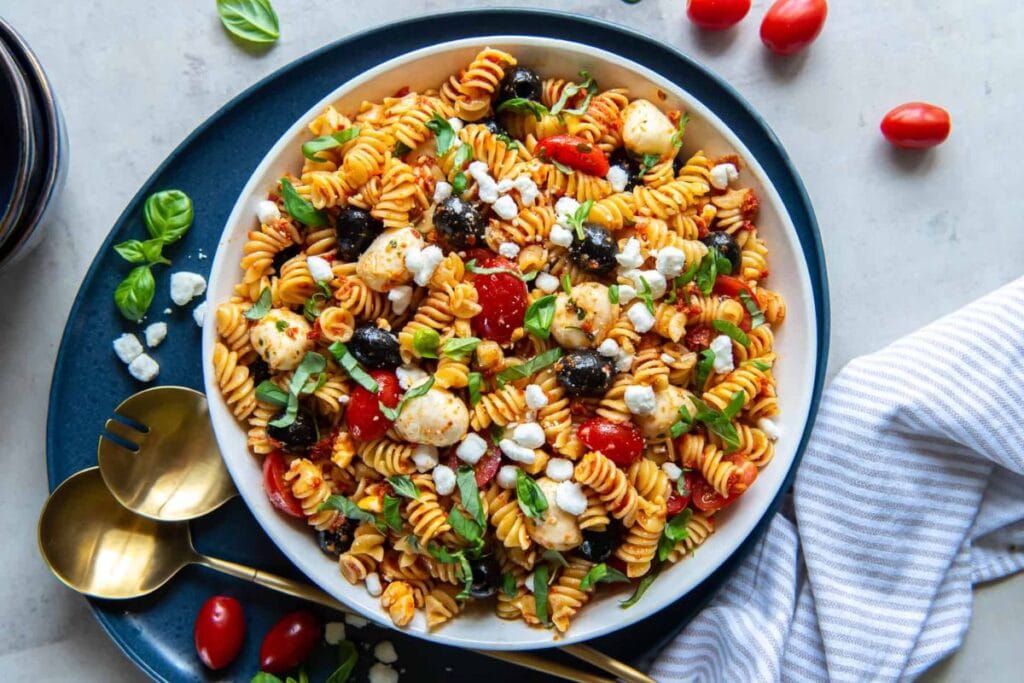 A bowl of pasta salad with rotini pasta, sun-dried tomatoes, mozzarella balls, olives, and feta cheese. Fresh basil is sprinkled on top.