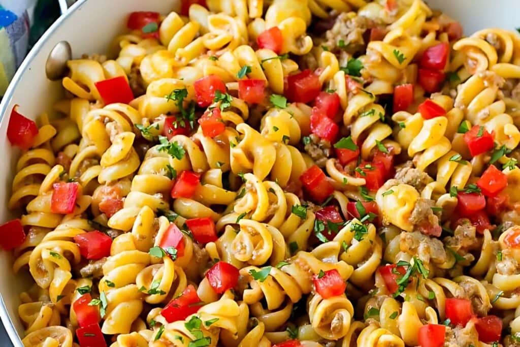 A close-up shot of taco pasta in a white skillet. The pasta is rotini, and it's mixed with ground beef, diced tomatoes, and chopped cilantro.