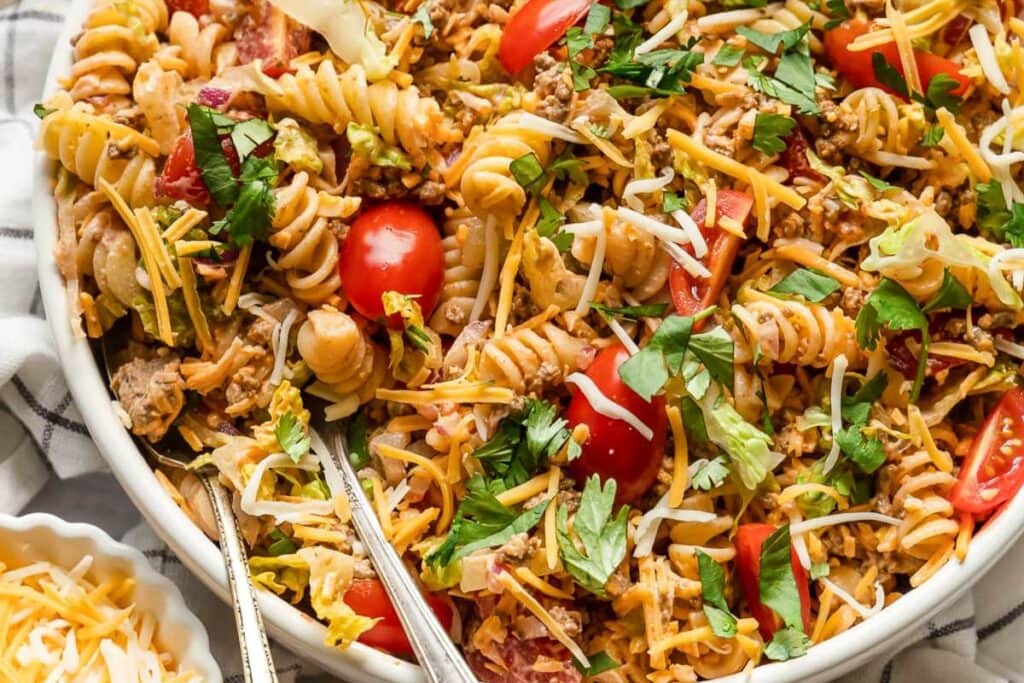 A bowl of pasta salad with rotini pasta, ground beef, shredded cheese, lettuce, tomatoes, and cilantro.