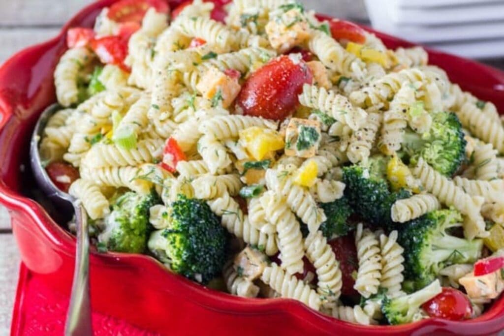A red bowl of pasta salad with rotini pasta, broccoli florets, cherry tomatoes, bell peppers, and cheese in a creamy dressing.