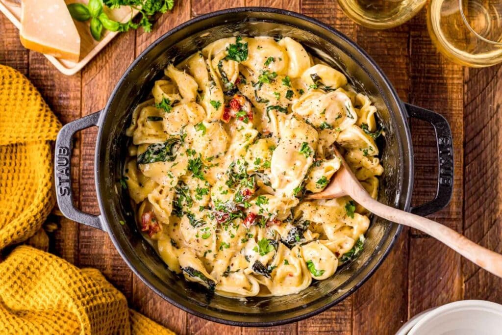 A black cast iron skillet of tortellini alla panna is shown from above. The tortellini is in a creamy sauce with spinach and sun-dried tomatoes, and it's topped with fresh parsley. A wooden spoon is in the skillet. A piece of Parmesan cheese and glasses of white wine are visible in the background.