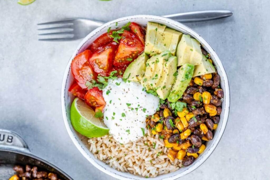 A vegetarian burrito bowl is shown from above. The bowl contains brown rice, black beans, corn, avocado, tomatoes, and a dollop of sour cream or yogurt. A lime wedge is on the side. A fork is visible in the upper left corner.
