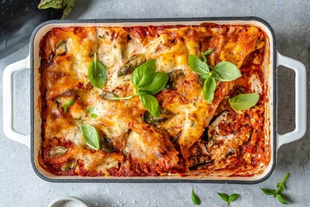 A rectangular baking dish of vegetarian eggplant lasagna is shown from above. The lasagna is topped with fresh basil leaves. A small bowl of salt is visible in the lower left corner.