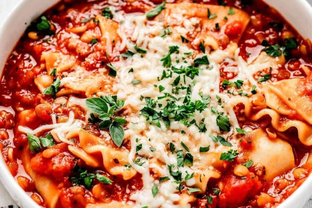 A bowl of vegetarian lasagna soup is shown from a close-up angle. The soup is a tomato-based broth with lasagna noodles, and it's topped with shredded cheese and fresh herbs.