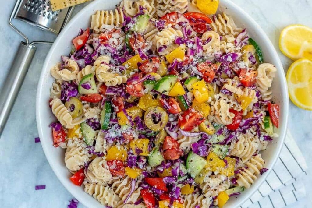 A white bowl of pasta salad with rotini pasta, red cabbage, cucumbers, bell peppers, tomatoes, and olives. Grated cheese is sprinkled on top.