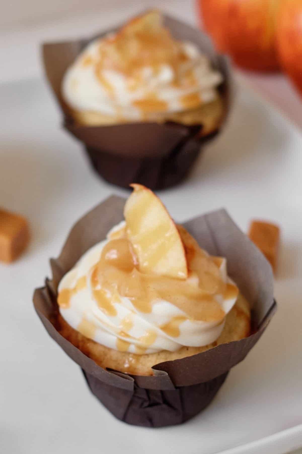 Caramel apple cupcakes in a brown cupcake liner on a white background with caramel candy and apples in the background