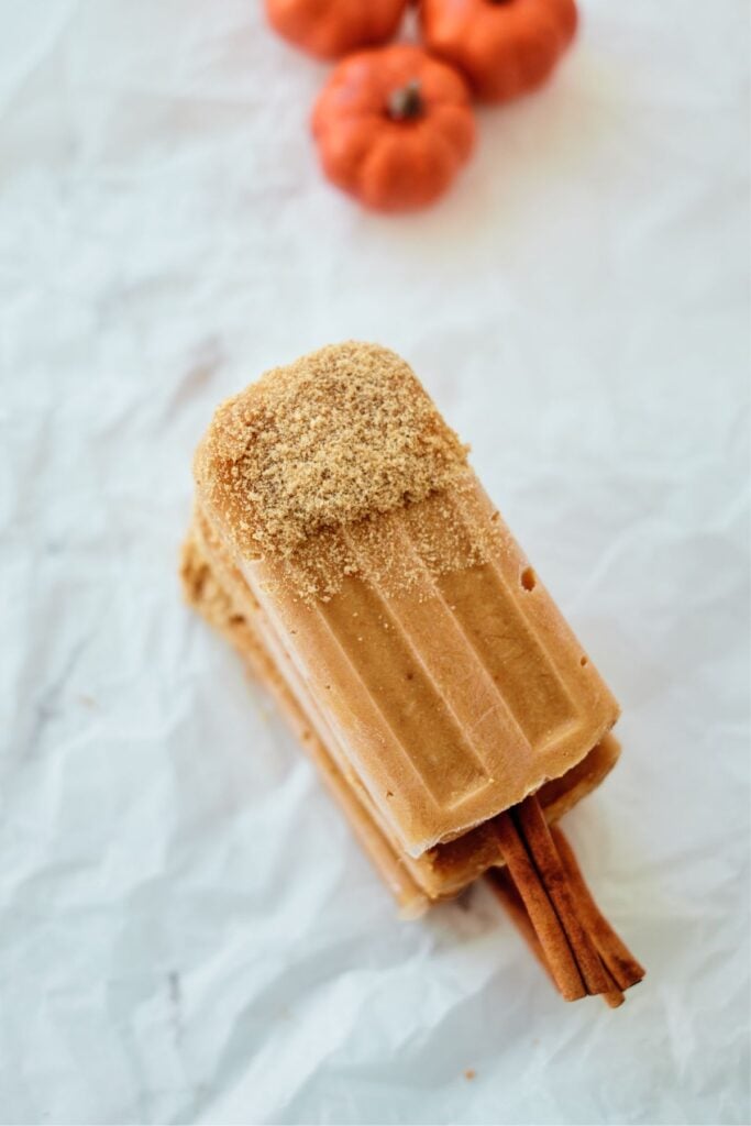 Pumpkin pie popsicles with cinnamon sticks and orange pumpkins in background.