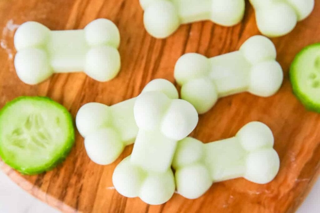 Light green, bone-shaped frozen dog treats and cucumber slices on a wooden board.