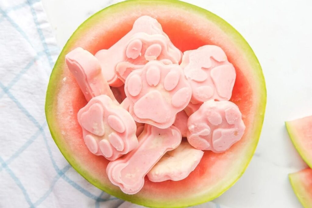 Pink, paw-print and bone-shaped frozen dog treats are in a watermelon rind bowl.