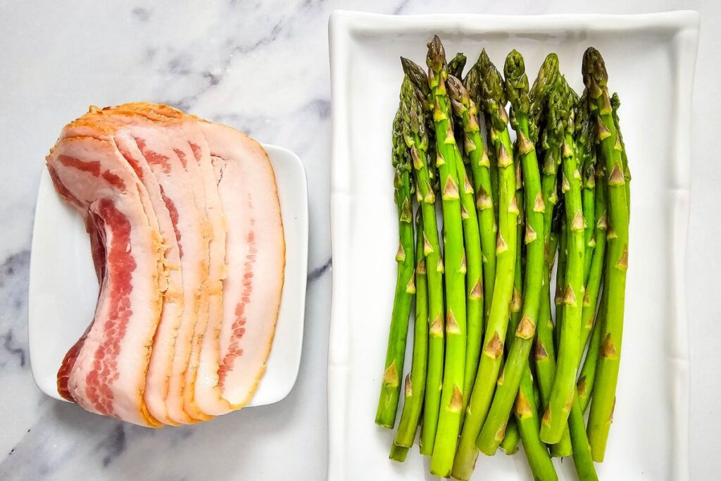 Raw asparagus and bacon arranged on separate white plates for prep.