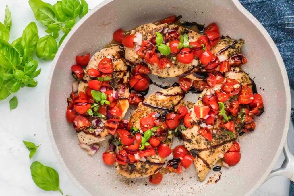 Bruschetta chicken in a white skillet topped with tomatoes and basil.