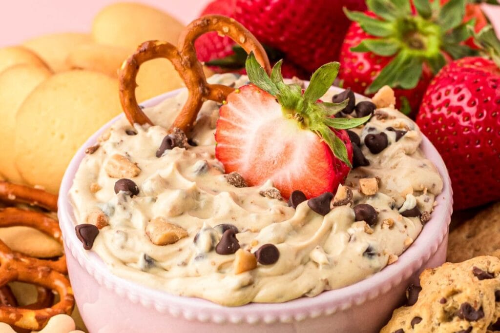 Chocolate chip cookie dough dip in a bowl, topped with a fresh strawberry and pretzel.