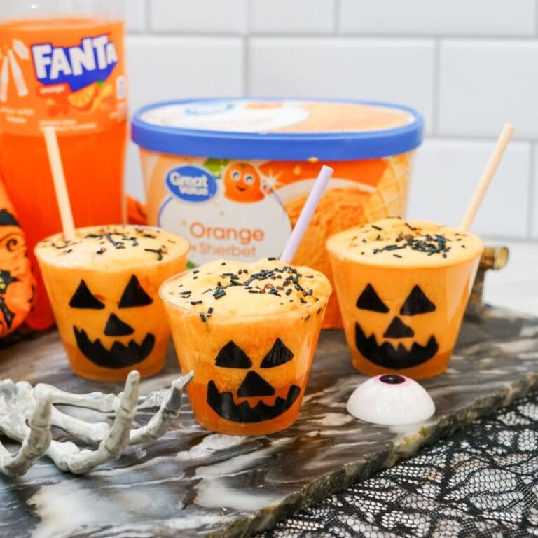 Fanta jack-o-lantern floats in orange cups with jack o lantern face. Orange sherbet and Fanta orange soda in the background.