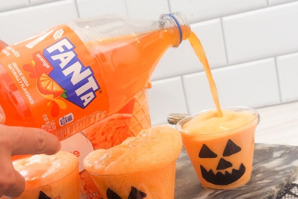 A hand pours orange Fanta into a clear plastic cup.
