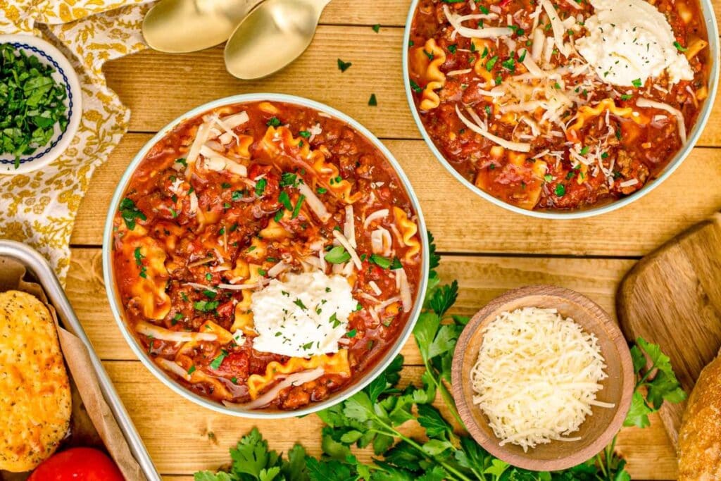 Two white bowls of lasagna soup topped with cheese and herbs.