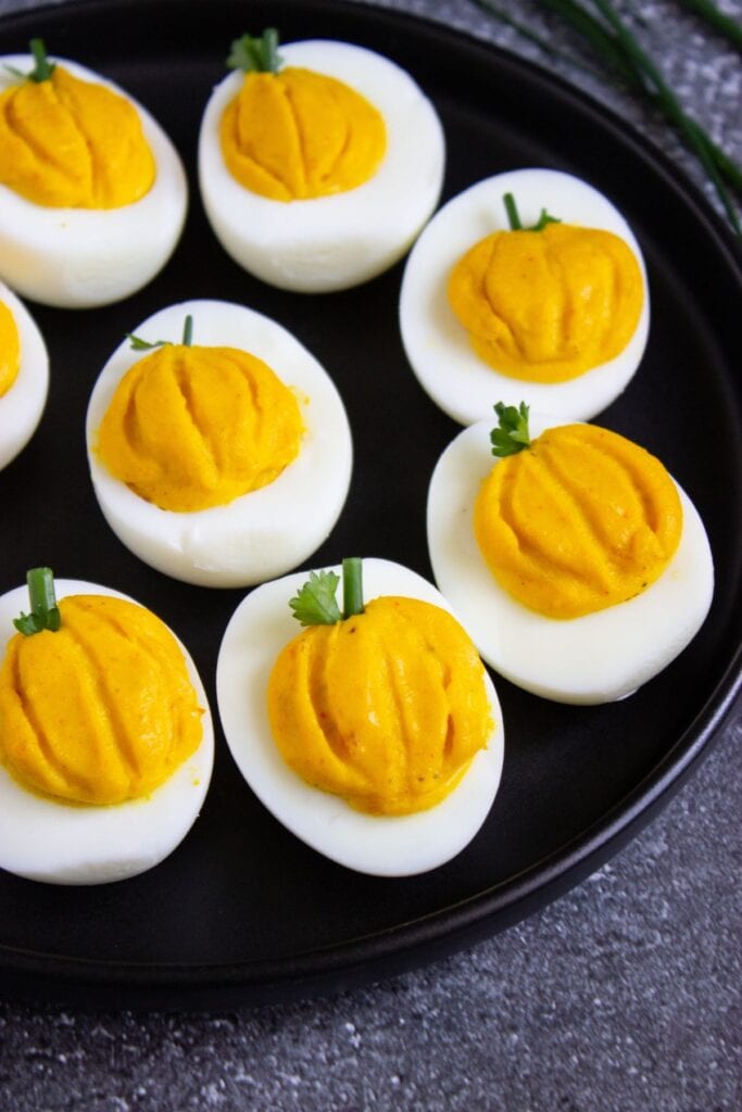 Pumpkin-shaped deviled eggs on a black plate, garnished with parsley and chive stems.
