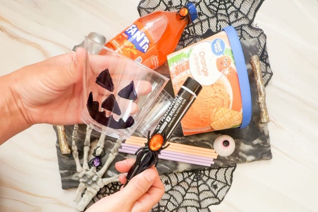 A pair of hands holds a clear plastic cup with a drawn jack-o'-lantern face.