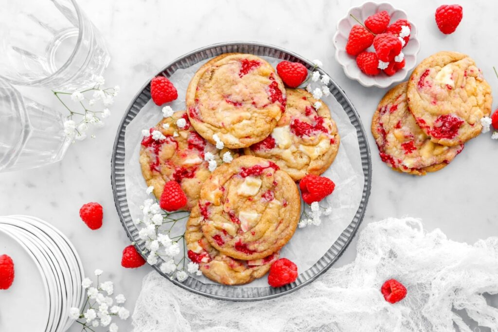 White chocolate chunk raspberry cookies on a plate with fresh raspberries and white flowers.