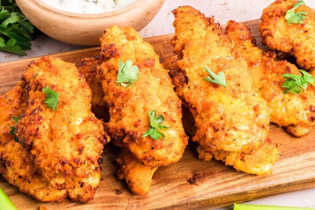 Buffalo chicken tenders on a wooden board, garnished with herbs and served with a dipping sauce.