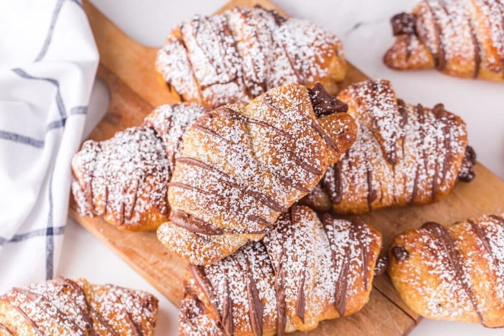Air Fryer Croissants with powdered sugar and chocolate drizzle on a wooden board.