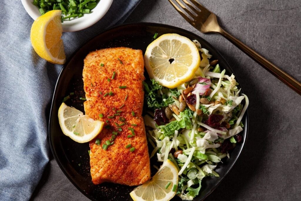 Air-fried salmon on a black plate with lemon, vegetables, and a fork on the side.