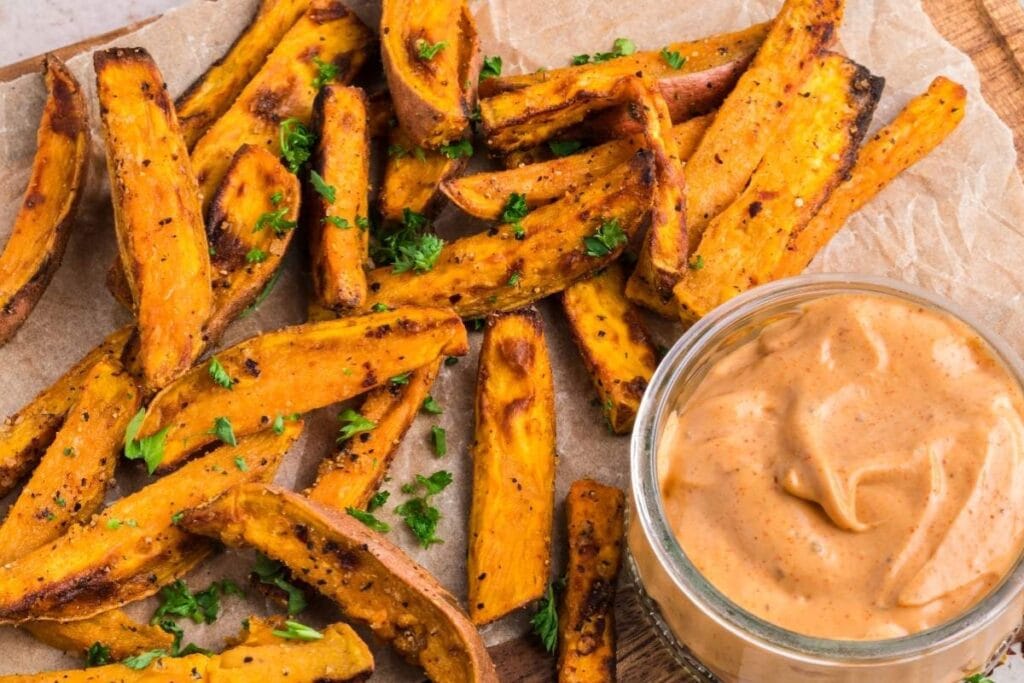 Air fryer sweet potato wedges with dip on parchment paper on a wooden board.