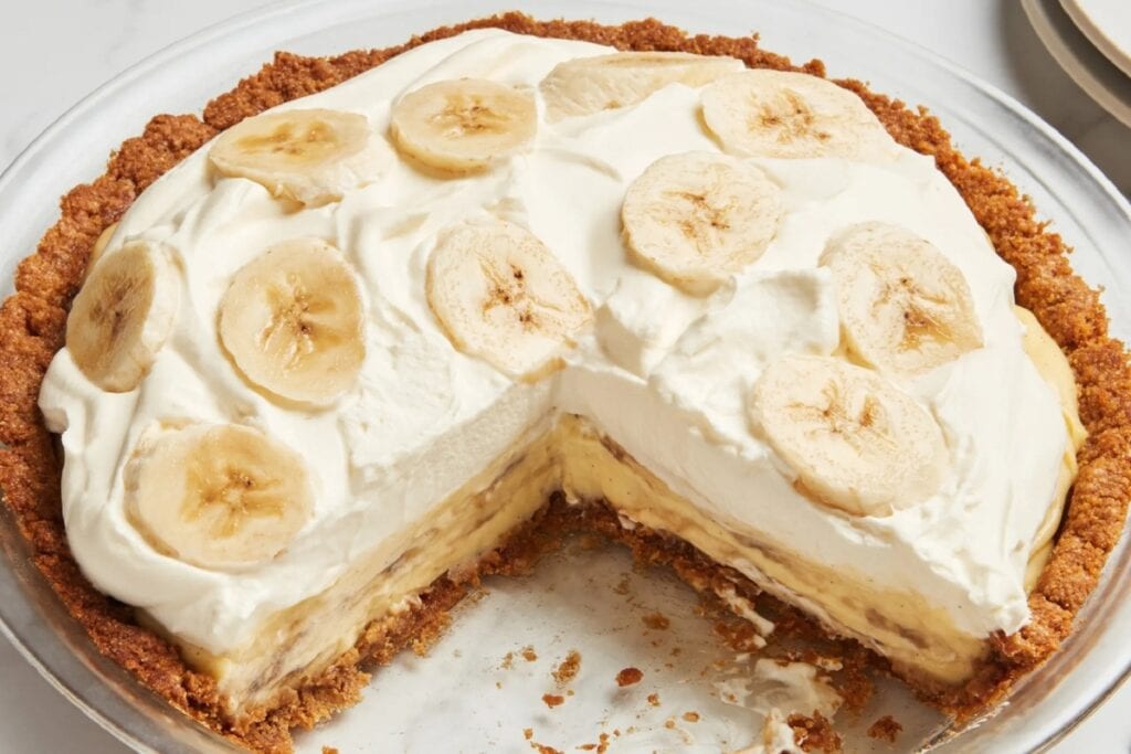 Banana cream pie in a clear glass pie dish with one slice removed.
