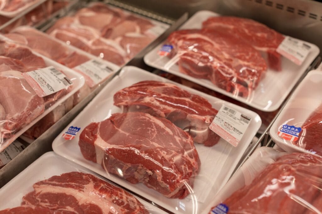Large packs of raw meat sealed in plastic, displayed in a grocery store fridge.