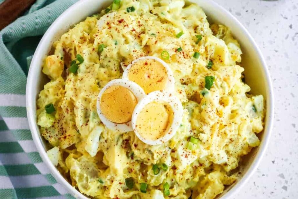 Classic Southern potato salad in a white bowl topped with sliced egg and green onions.