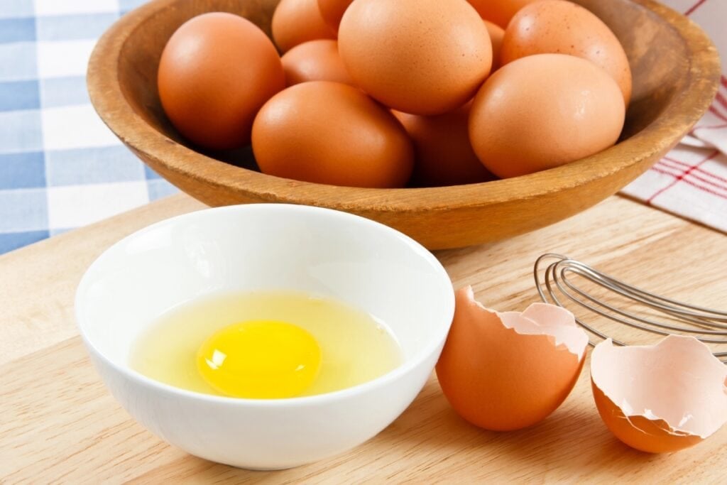 Brown eggs in a wooden bowl, with one raw egg cracked into a white bowl.
