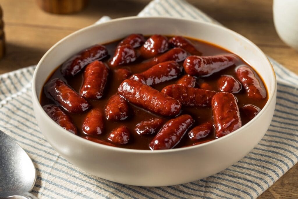 Lil’ smokies simmered in barbecue sauce in a white bowl.