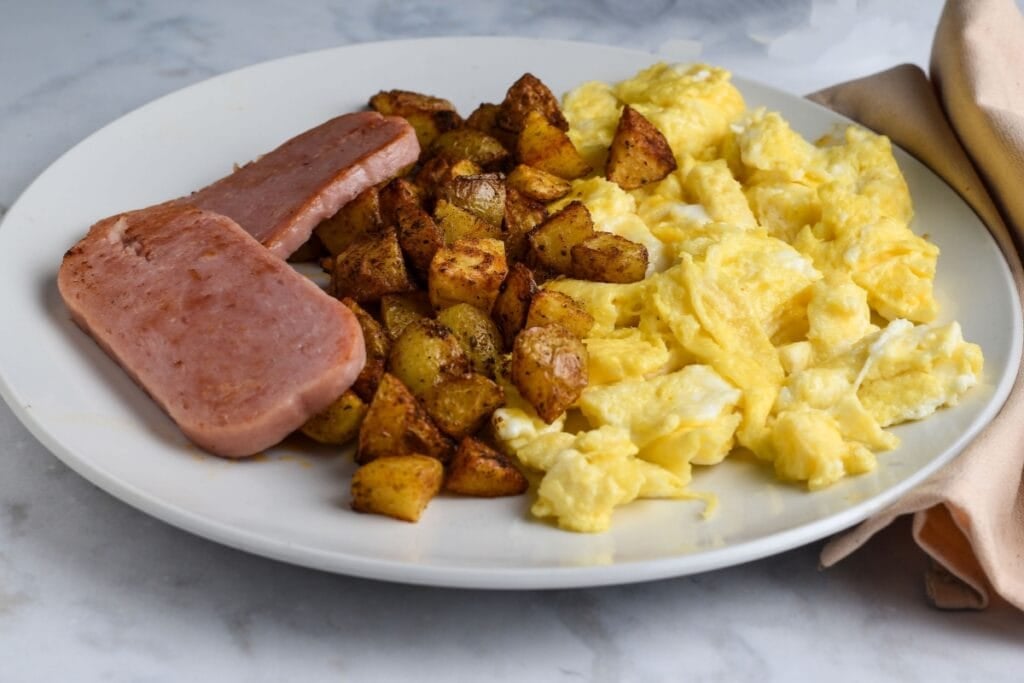 Fried spam, potatoes, and scrambled eggs on a white plate.
