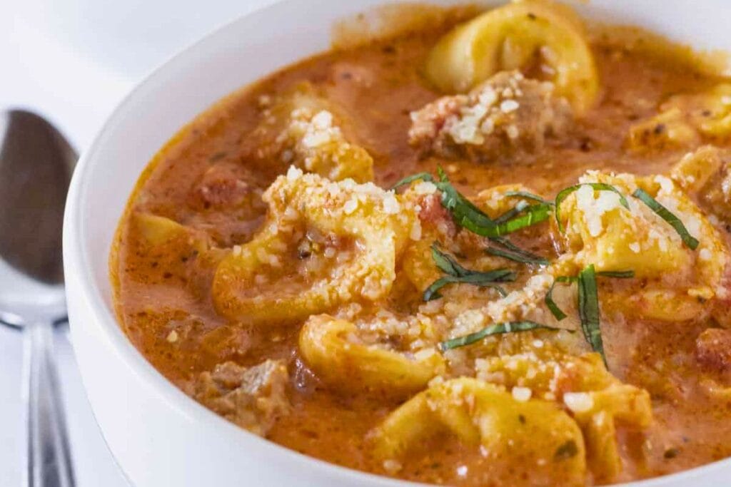 Close-up of sausage tortellini soup with basil and parmesan in a white bowl.