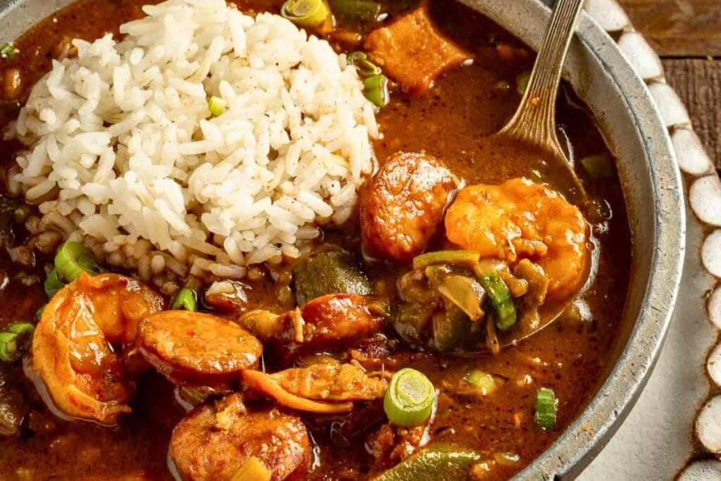 Gumbo with shrimp, sausage, and rice in a bowl, being served with a spoon.