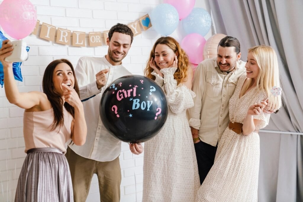 Young excited couple blowing up surprise balloon during gender reveal party indoors.